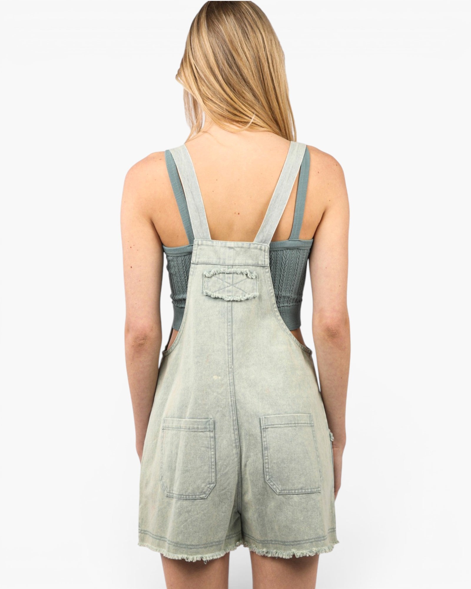 A woman shown from the back wearing a light sage green denim overall romper with frayed hems and wide shoulder straps, set against a plain white studio background.