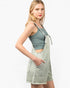 A blonde model wearing a sage green ribbed tank top and matching sage green denim overall shorts with a tie waist and front pockets, posed against a solid white background.