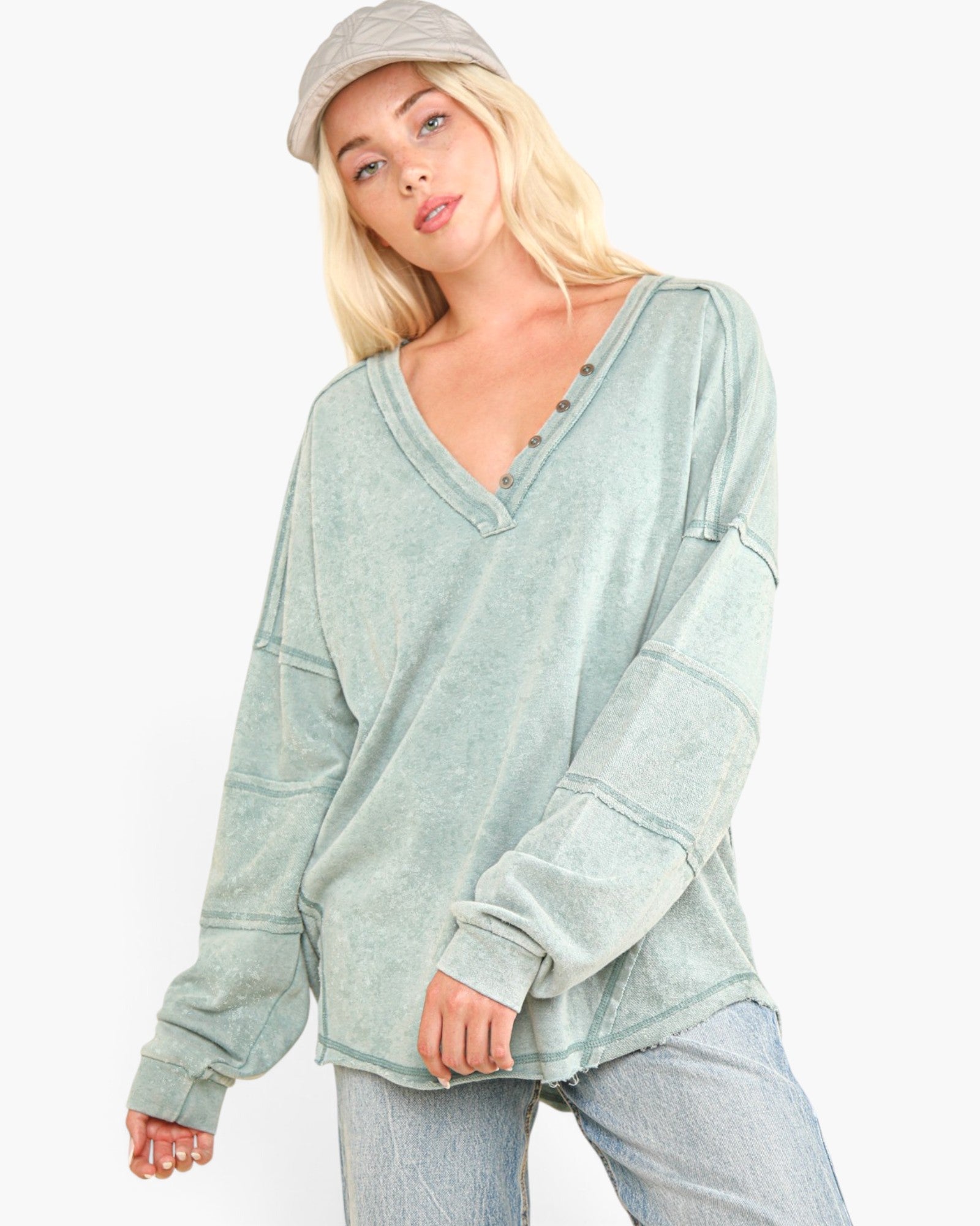 A woman wearing a sage green, long-sleeve V-neck henley top with a washed, distressed texture, paired with light-wash jeans and a beige cap, against a plain white background.