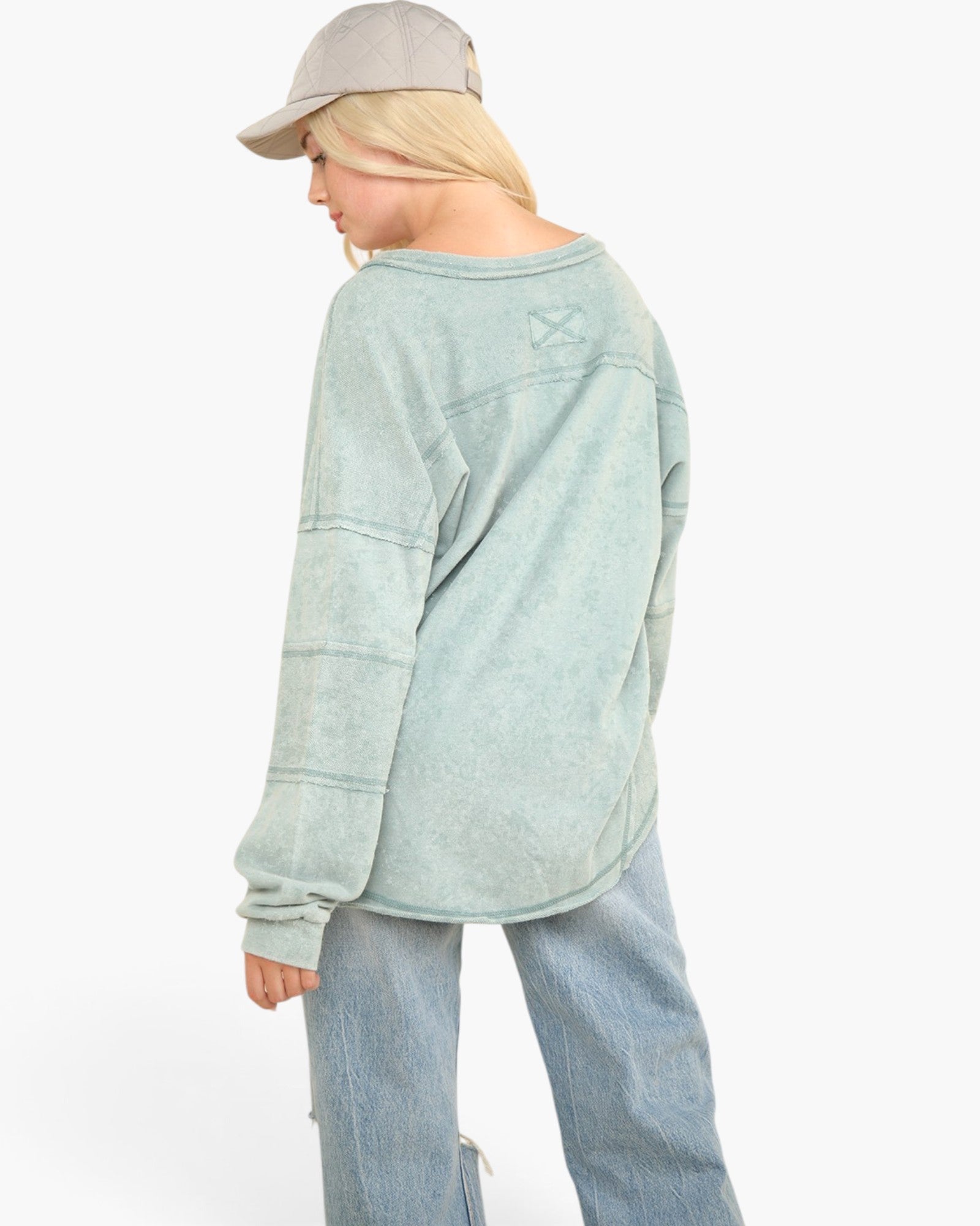 A model shown from the back wearing a light sage green long-sleeve sweatshirt, light blue jeans, and a gray baseball cap against a plain white background.