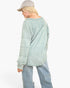 A model shown from the back wearing a light sage green long-sleeve sweatshirt, light blue jeans, and a gray baseball cap against a plain white background.