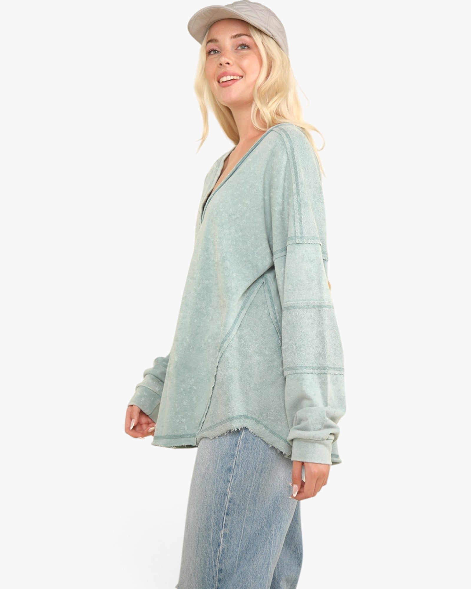 A woman wearing a sage green, long-sleeve, V-neck knit top with a raw, frayed hem and side slits, paired with light-wash jeans and a gray cap, photographed against a solid white background.