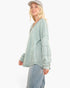A woman wearing a sage green, long-sleeve, V-neck knit top with a raw, frayed hem and side slits, paired with light-wash jeans and a gray cap, photographed against a solid white background.