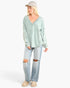 A woman modeling a light sage green long-sleeve V-neck top with distressed light-wash jeans, beige sneakers, and a beige baseball cap against a plain white studio background.