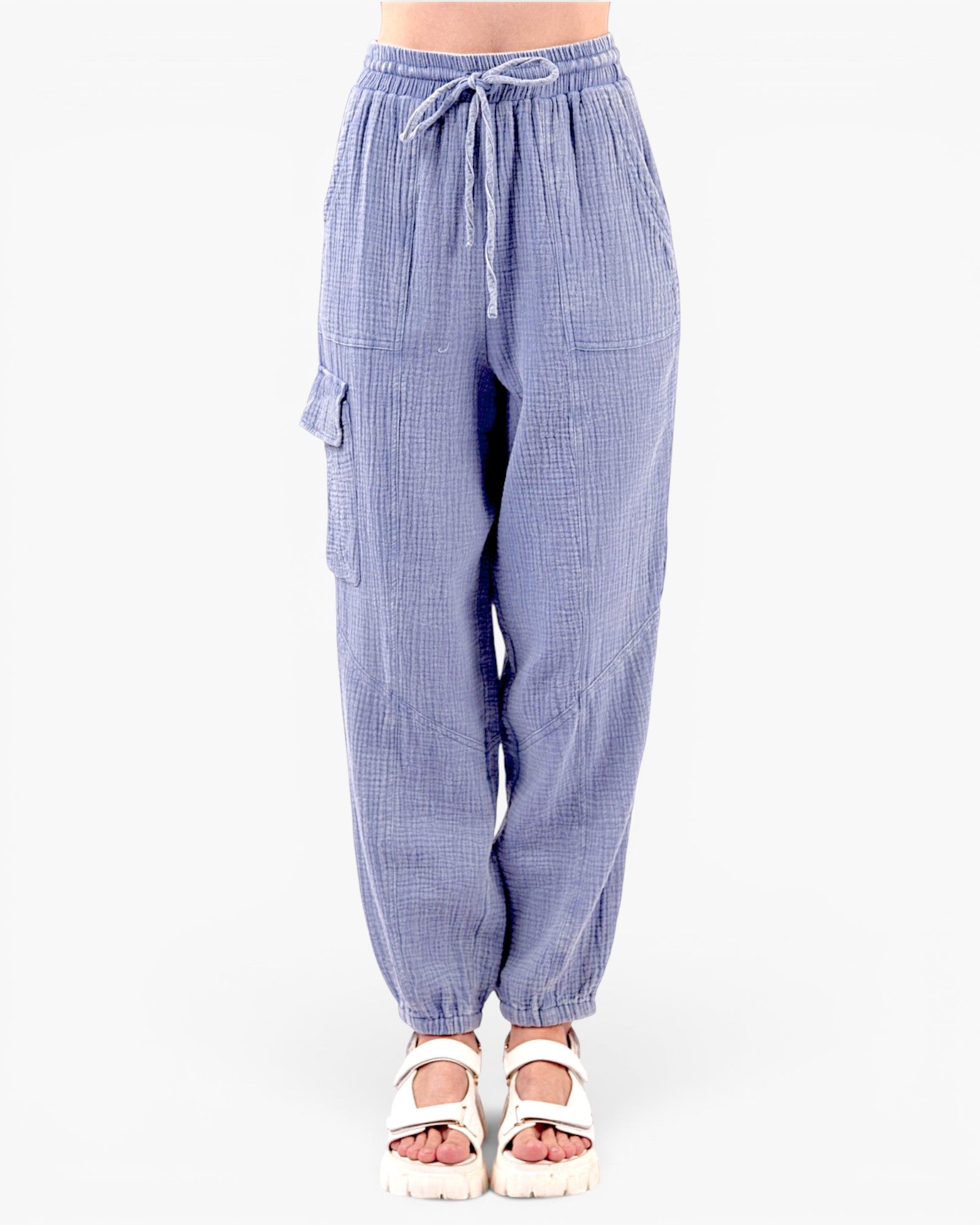 A person wearing loose, light blue gingham cargo-style pants with an elastic drawstring waist and side flap pockets, paired with white platform sandals, against a plain white studio background.