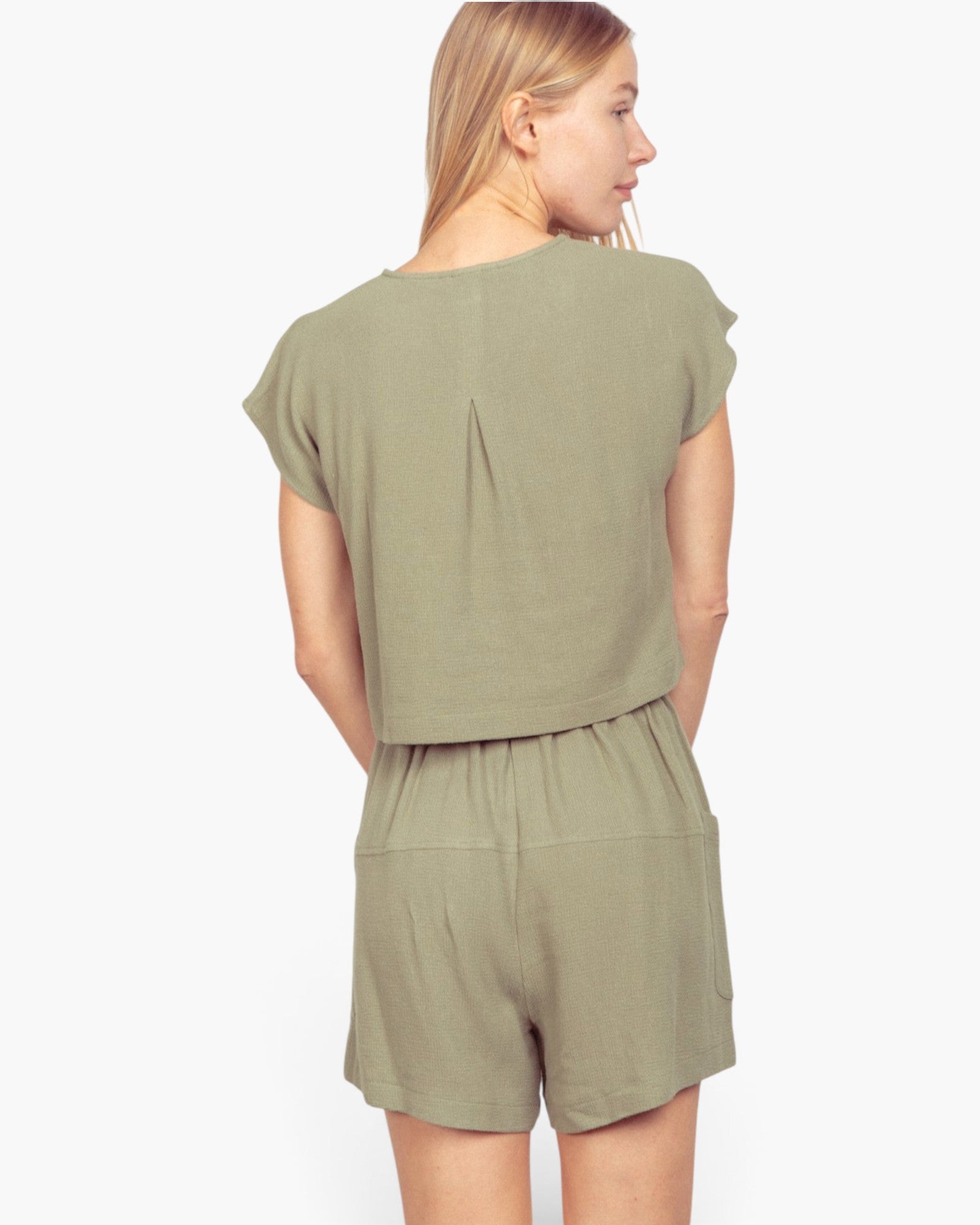 A woman shown from the back wearing a matching sage green short-sleeve top and shorts set against a plain light gray studio background.