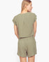 A woman shown from the back wearing a matching sage green short-sleeve top and shorts set against a plain light gray studio background.