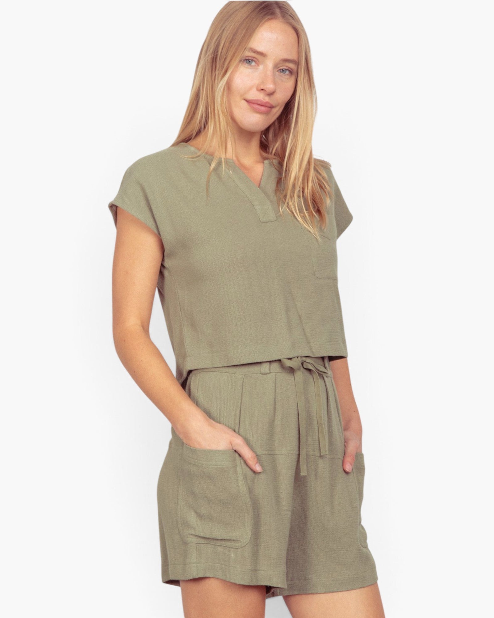 A woman modeling a matching two-piece lounge set in olive green, featuring a short-sleeve top with a split neckline and matching drawstring shorts, set against a plain light gray background.