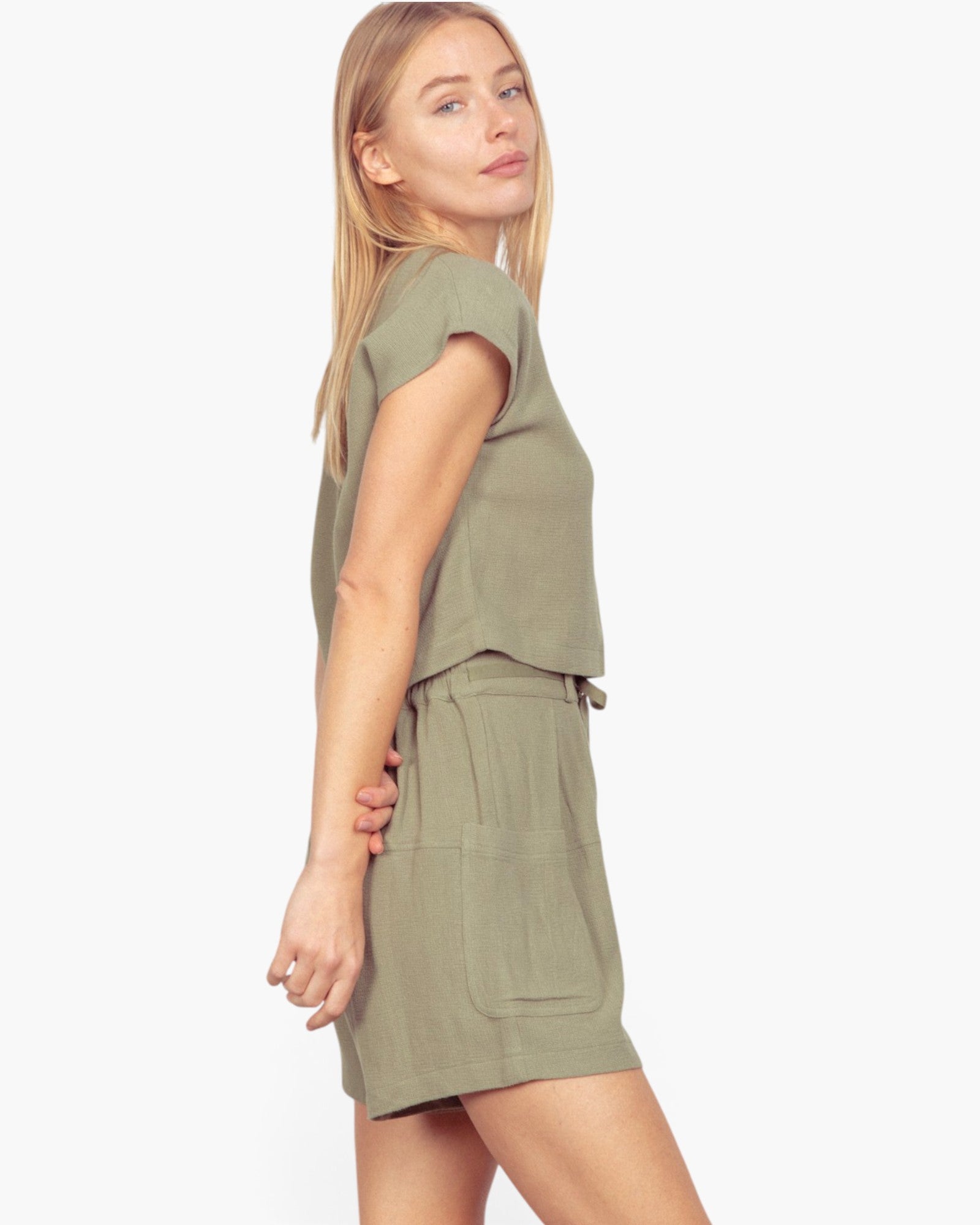 A woman modeling a matching olive green short-sleeve crop top and high-waisted mini skirt set against a plain white studio background.