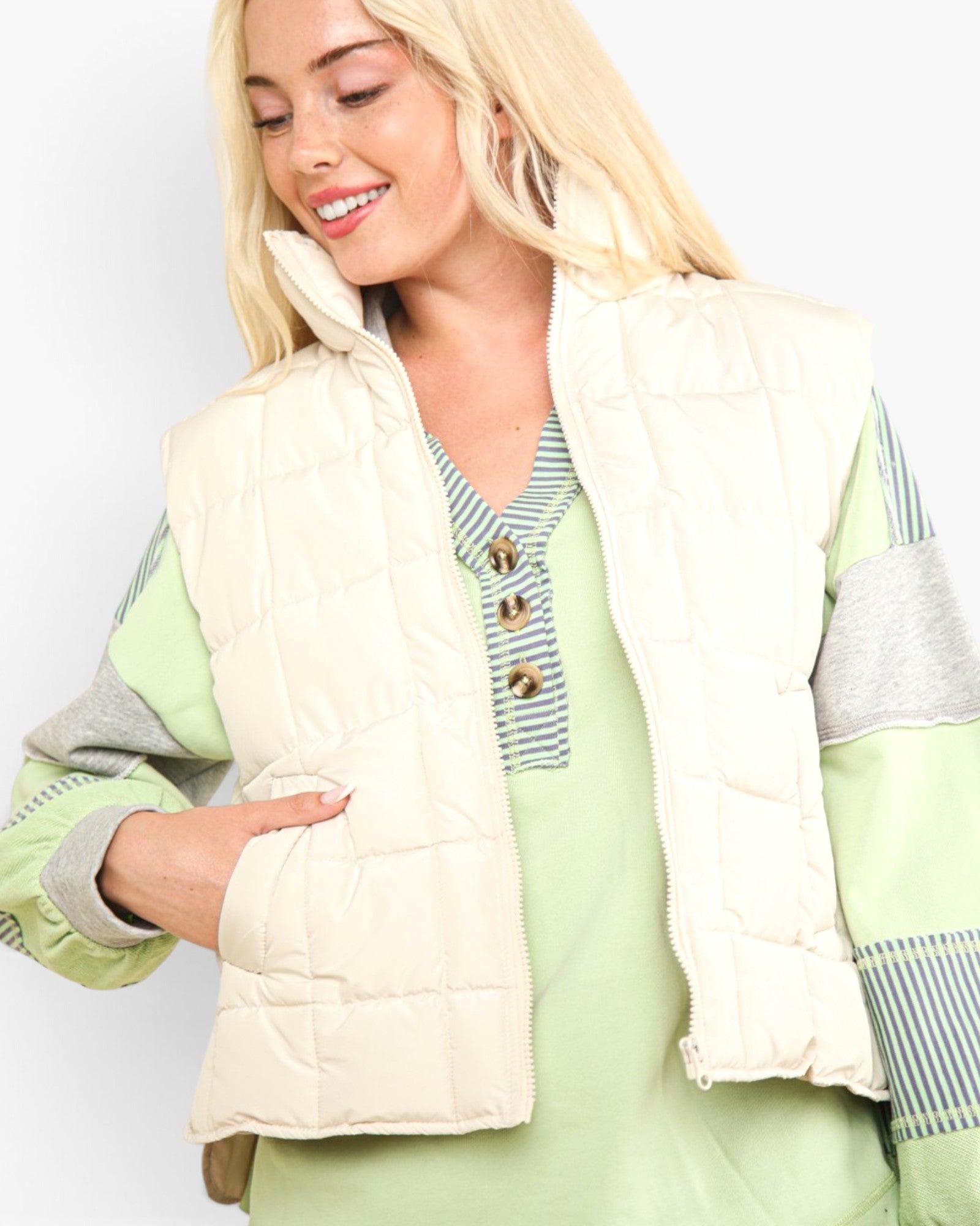 A blonde woman wearing a cream quilted sleeveless puffer vest over a light green and gray color-blocked knit top, photographed against a plain light gray studio background.