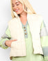 A blonde woman wearing a cream quilted sleeveless puffer vest over a light green and gray color-blocked knit top, photographed against a plain light gray studio background.