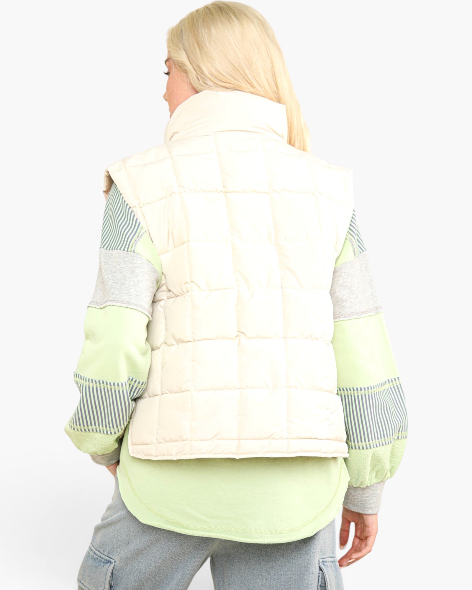 A woman shown from the back wearing a cream quilted puffer vest over a light green and gray color-blocked long-sleeve shirt, paired with gray pants, against a plain white background.