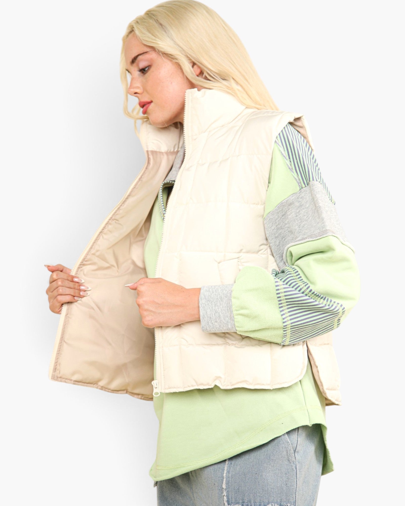 A blonde woman wearing a cream quilted puffer vest over a sage green and gray color-blocked long-sleeve top, paired with light blue jeans, shown against a plain white background.