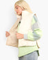 A blonde woman wearing a cream quilted puffer vest over a sage green and gray color-blocked long-sleeve top, paired with light blue jeans, shown against a plain white background.