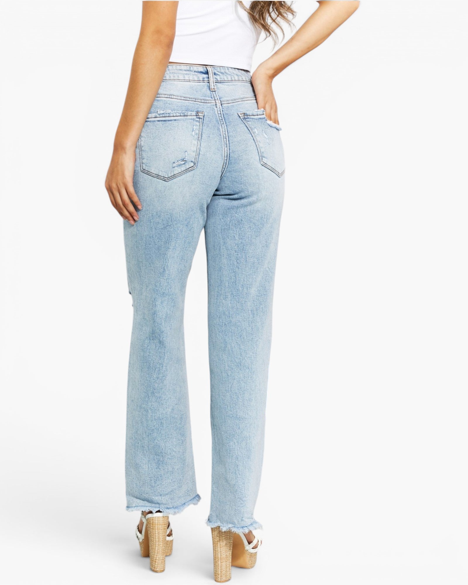 A model shown from the back wearing light-wash blue denim jeans with a straight-to-wide leg fit, cuffed hems, and distressed details on the back pockets, paired with a white top and beige platform sandals.