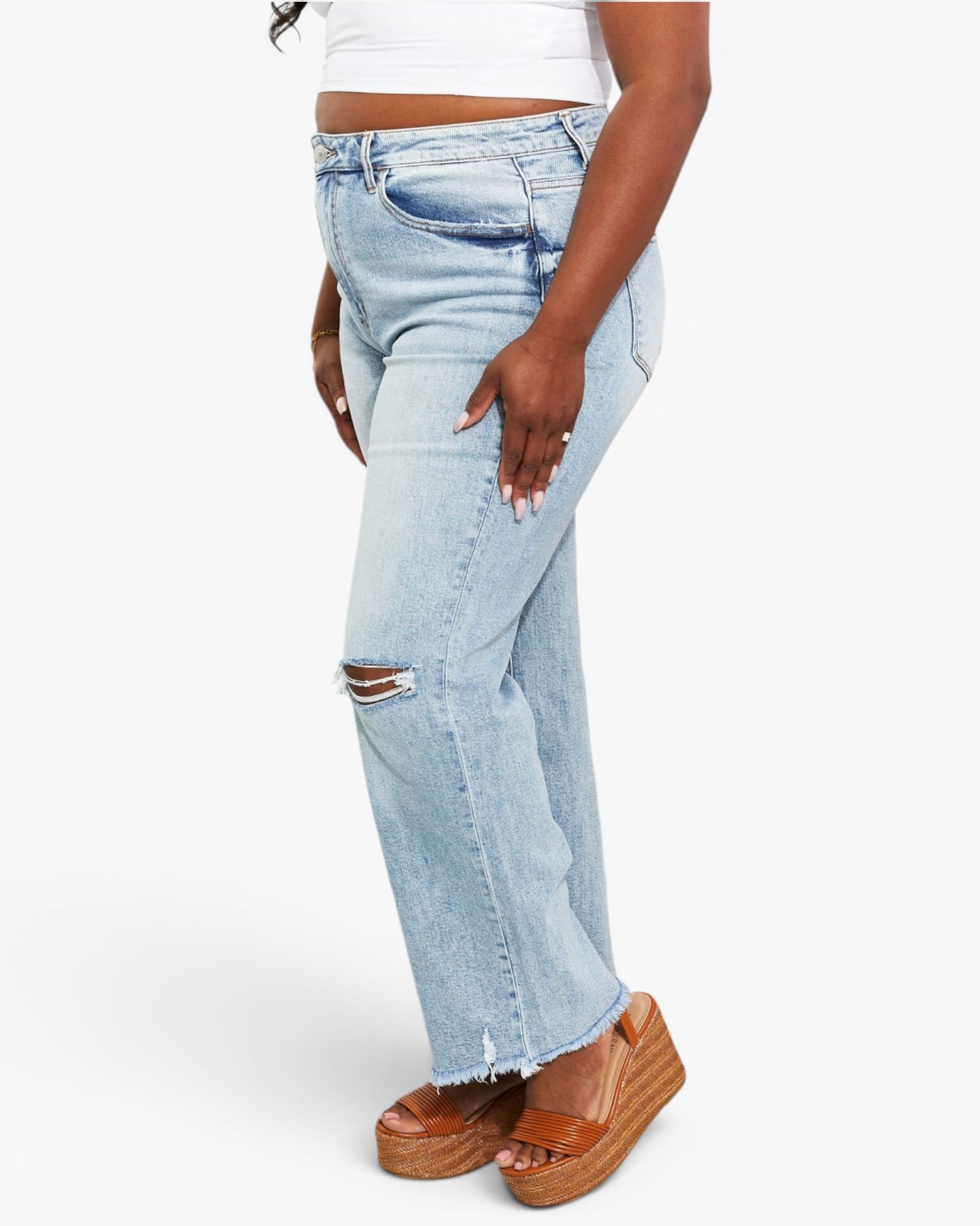 A model wearing light-wash blue distressed jeans with a raw frayed hem, paired with brown platform sandals, shown from the waist down against a plain white studio background.