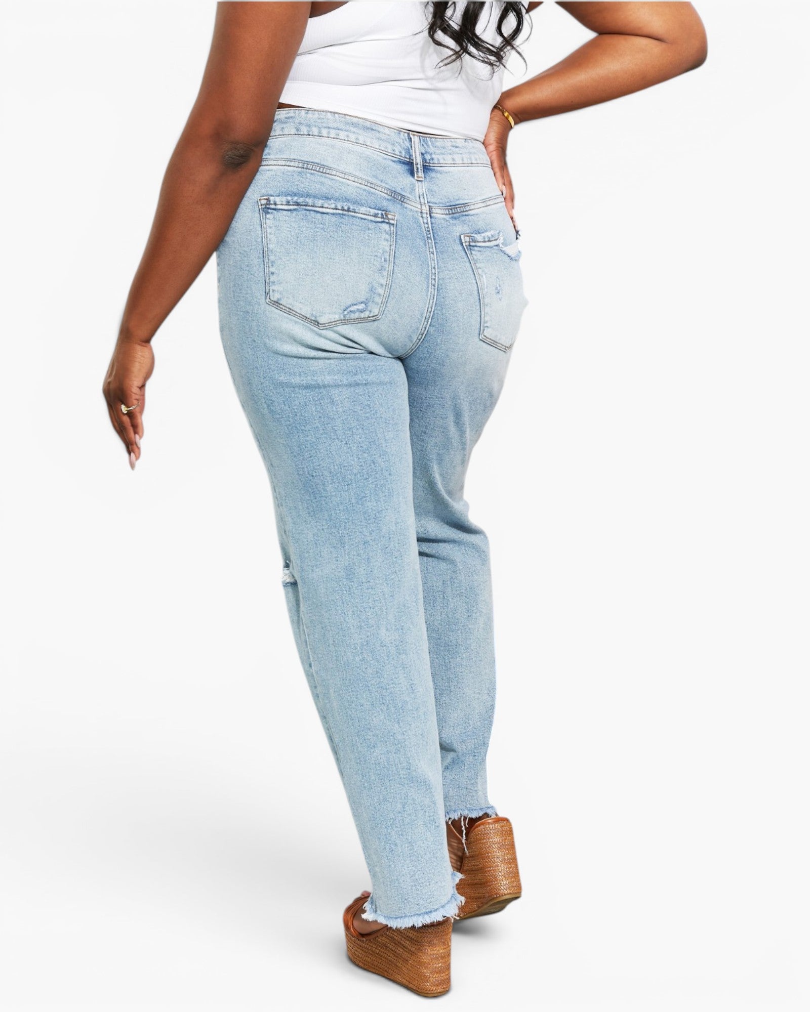 A person shown from the back wearing light-wash blue jeans with a white top and brown platform shoes against a plain light gray studio background.