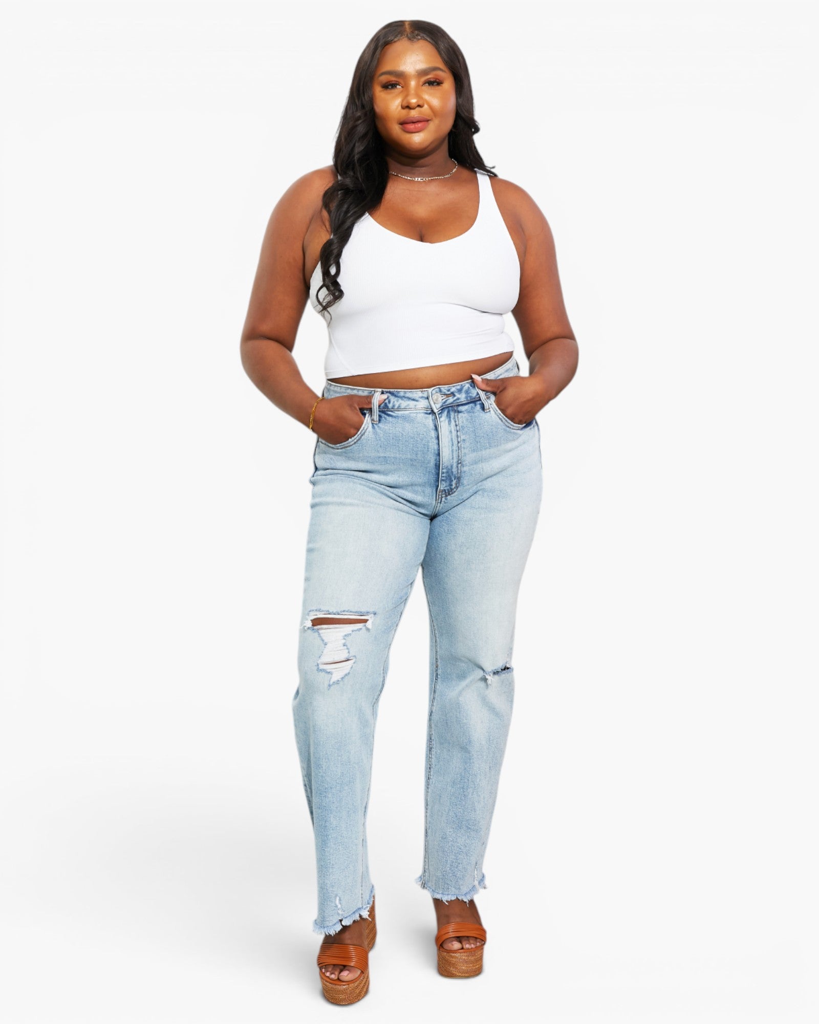 A woman modeling light-wash blue distressed jeans with a white crop top and brown platform sandals against a plain white studio background.