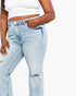 A close-up of a model wearing light-wash blue denim jeans with a white cropped tank top, showing the front pockets and a distressed rip on the thigh.