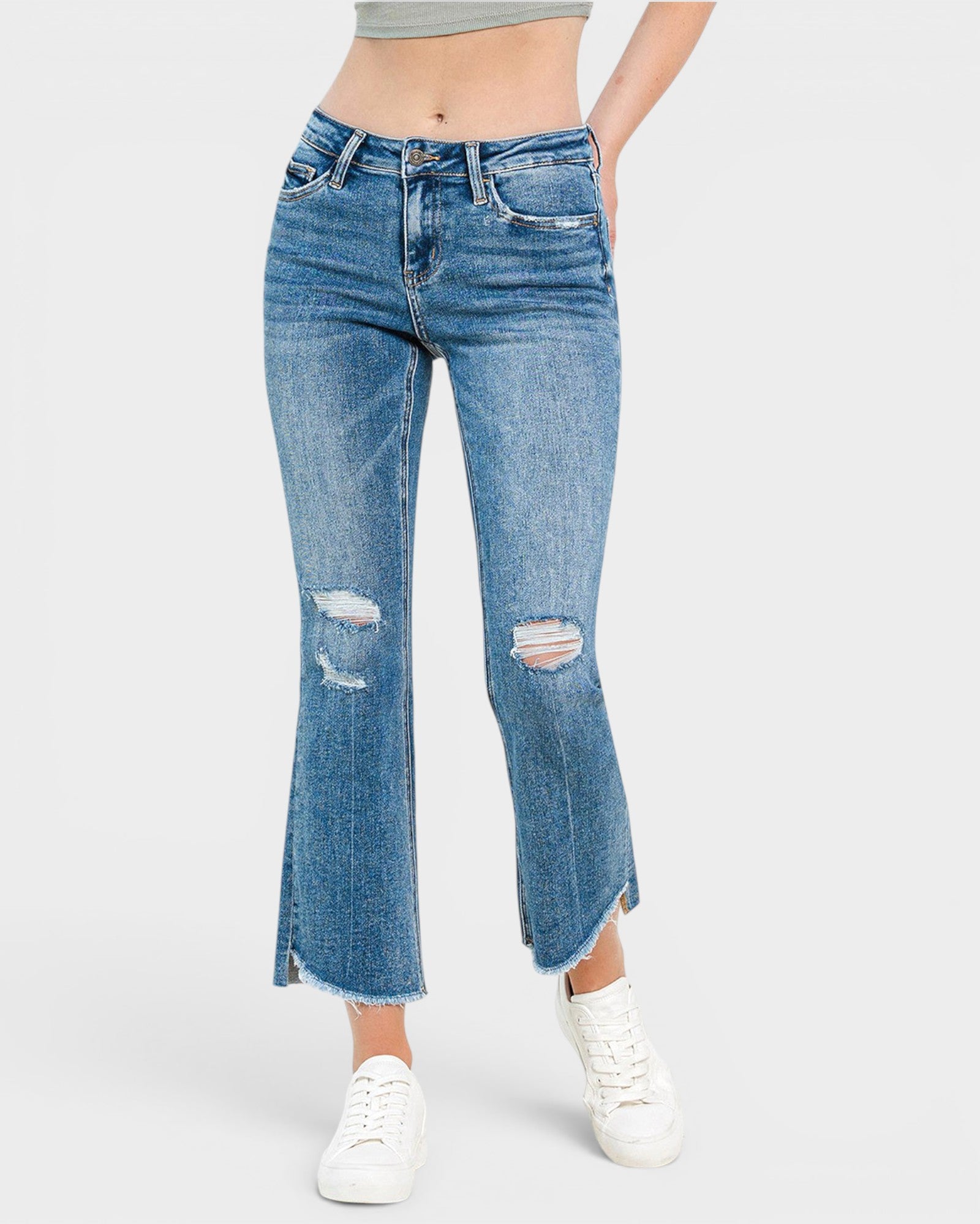 A model wearing a pair of medium-wash blue denim flare jeans with distressed rips on the thighs and hem, styled with white sneakers against a light gray background.