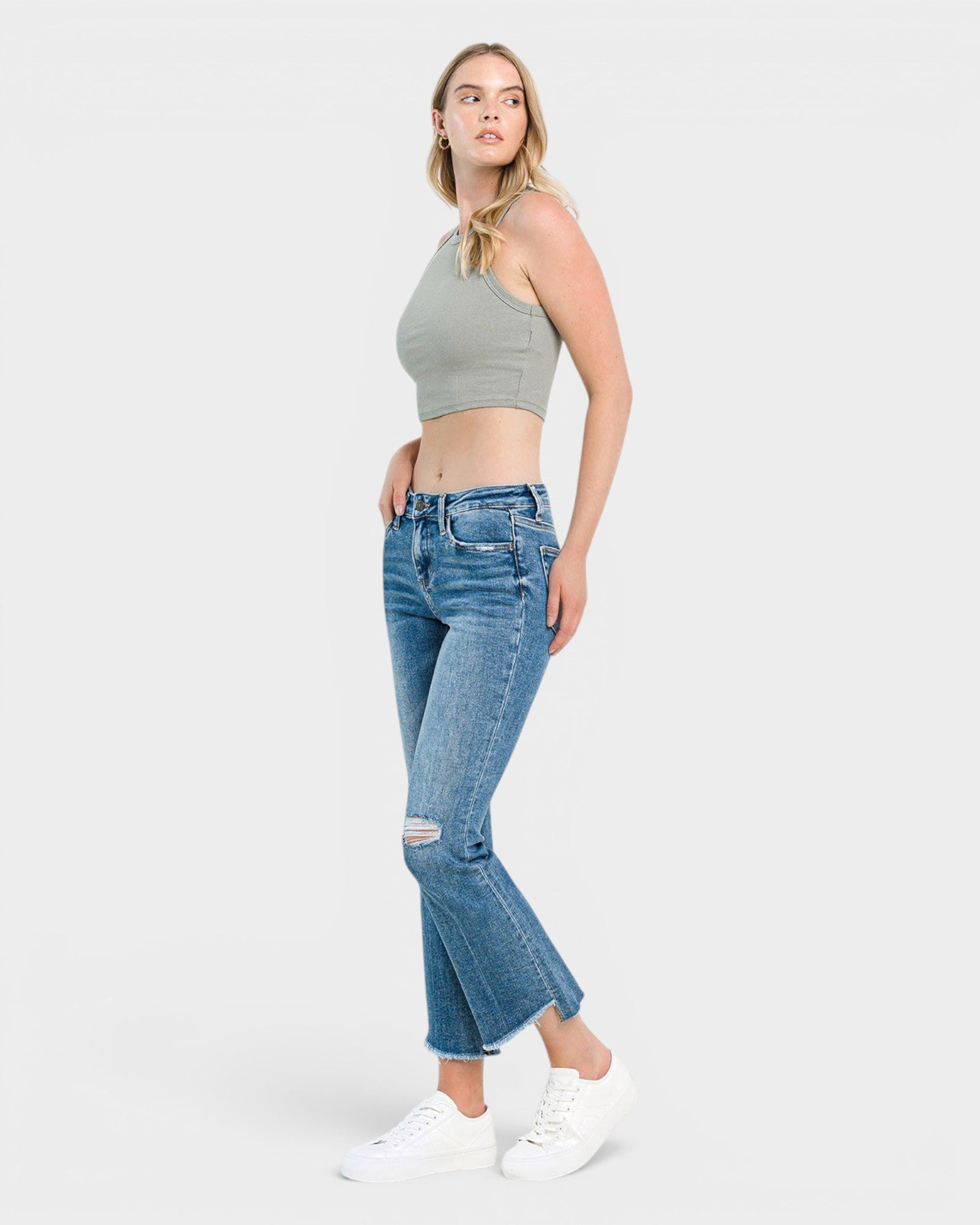 A woman modeling a light gray ribbed crop top and blue cropped flare jeans with distressed knees, paired with white sneakers, against a light gray studio background.