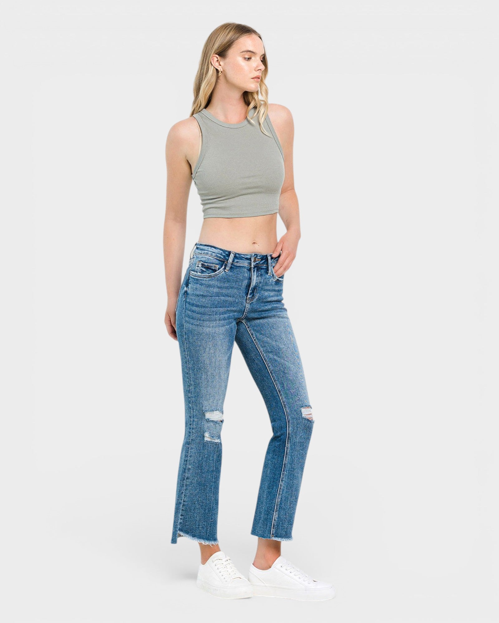 A woman modeling a pair of high-waisted, distressed blue jeans with a slight flare, paired with a gray ribbed crop top and white sneakers, set against a plain light gray studio background.