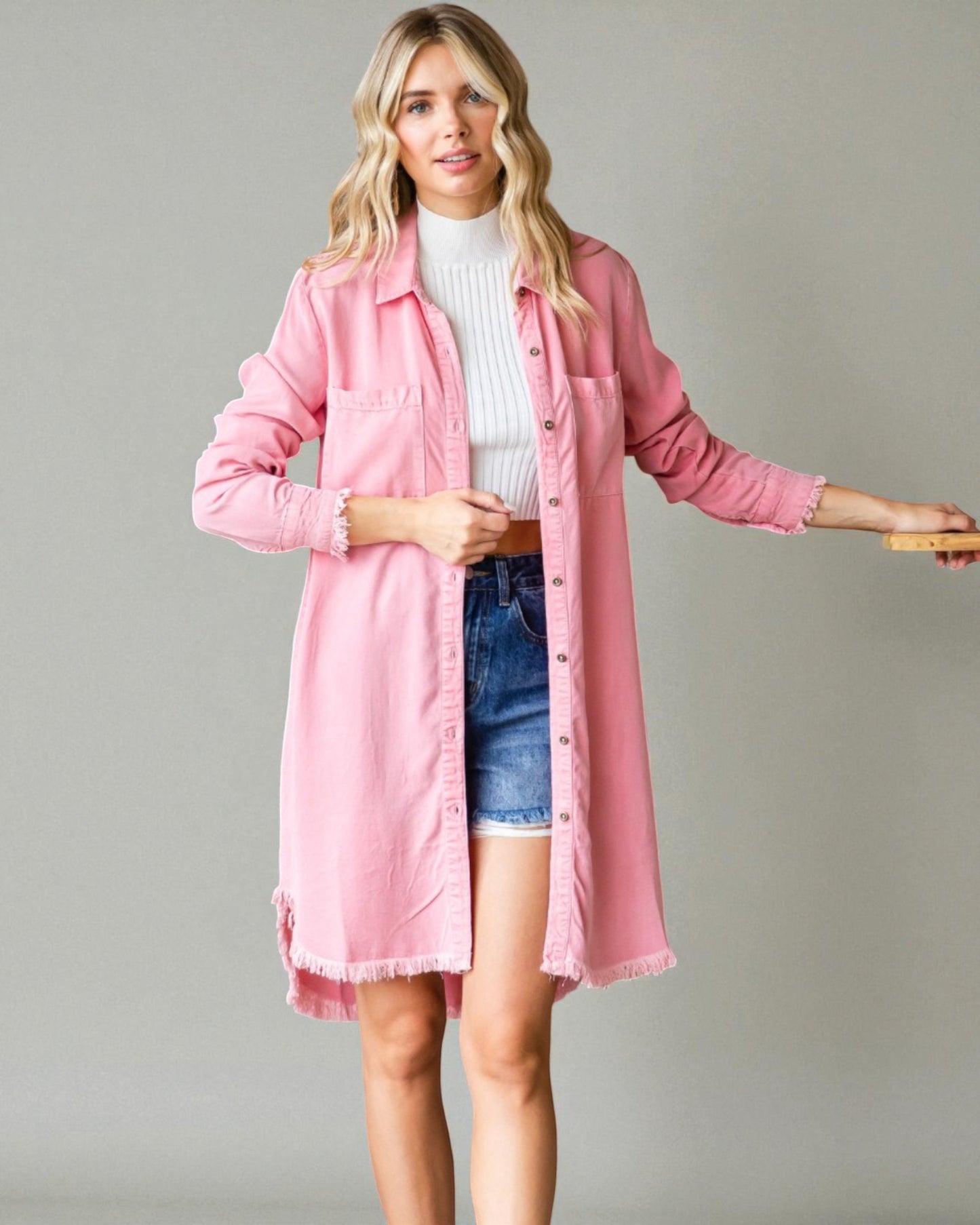 Woman wearing a pink jacket over a white sweater and denim shorts on a gray background