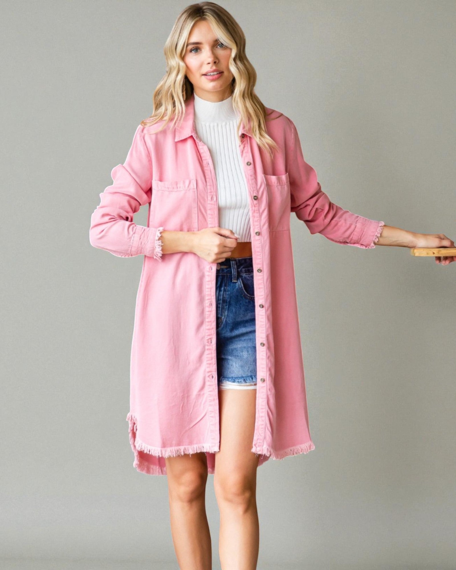 Woman wearing a pink jacket over a white sweater and denim shorts on a gray background