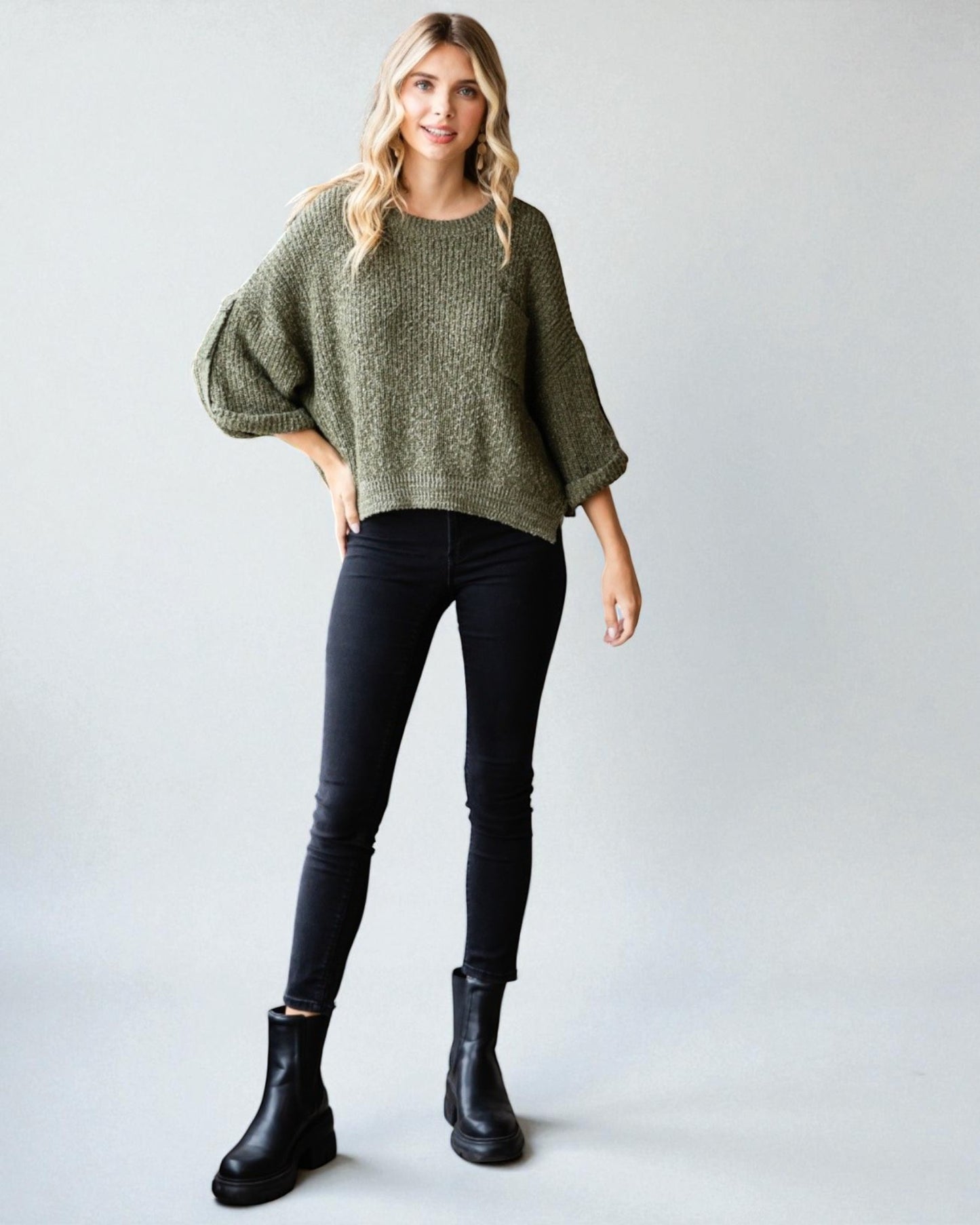 Woman wearing a green sweater, black pants, and black boots on a plain background