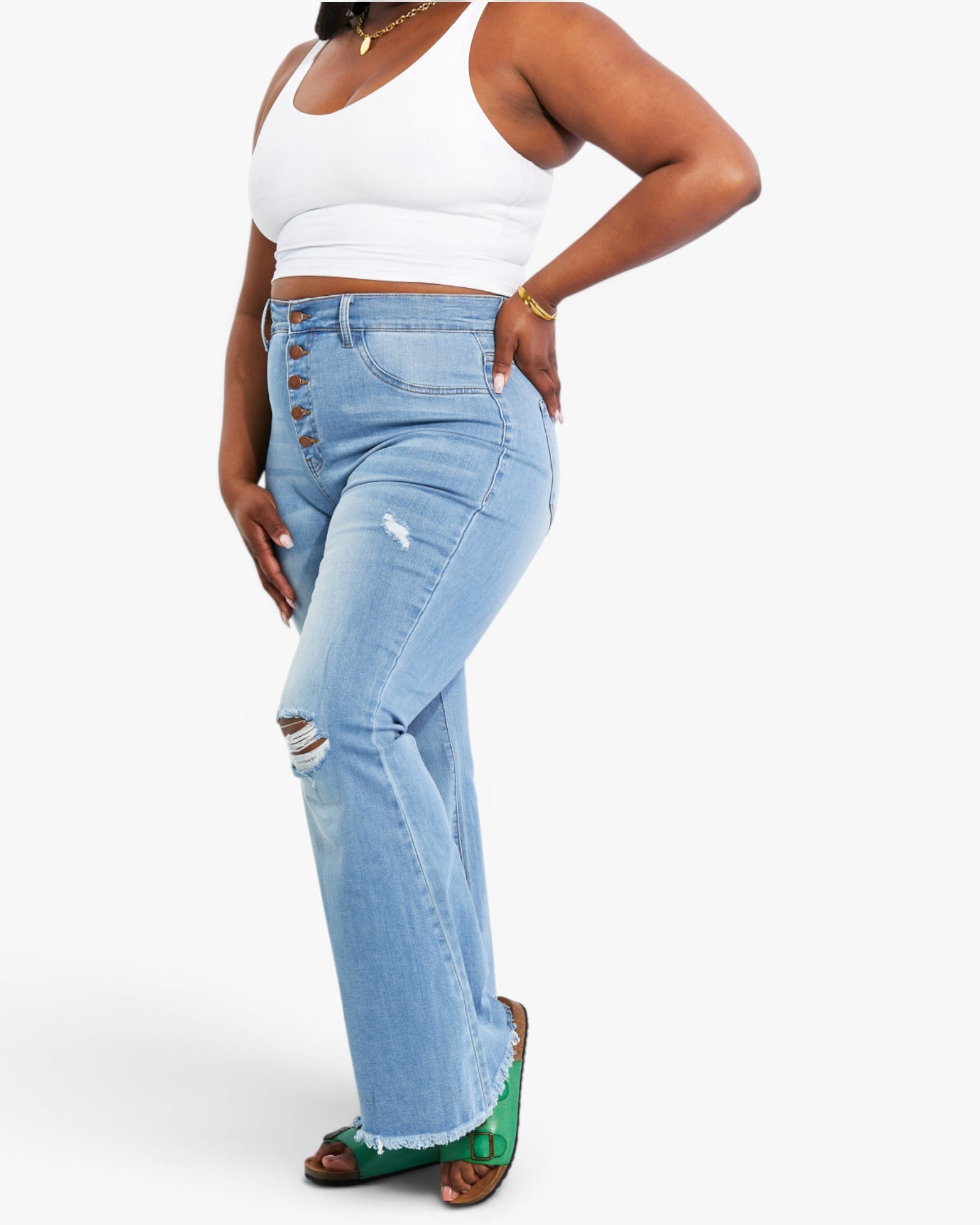 A model wearing high-waisted, light blue denim jeans with a button-fly front, distressed details, and a frayed hem, paired with a white crop top and green sandals.