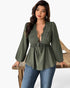 A woman models an olive green long-sleeve blouse with a lace V-neck bodice and drawstring waist, paired with blue jeans, standing behind a rustic wooden table against a white studio background.