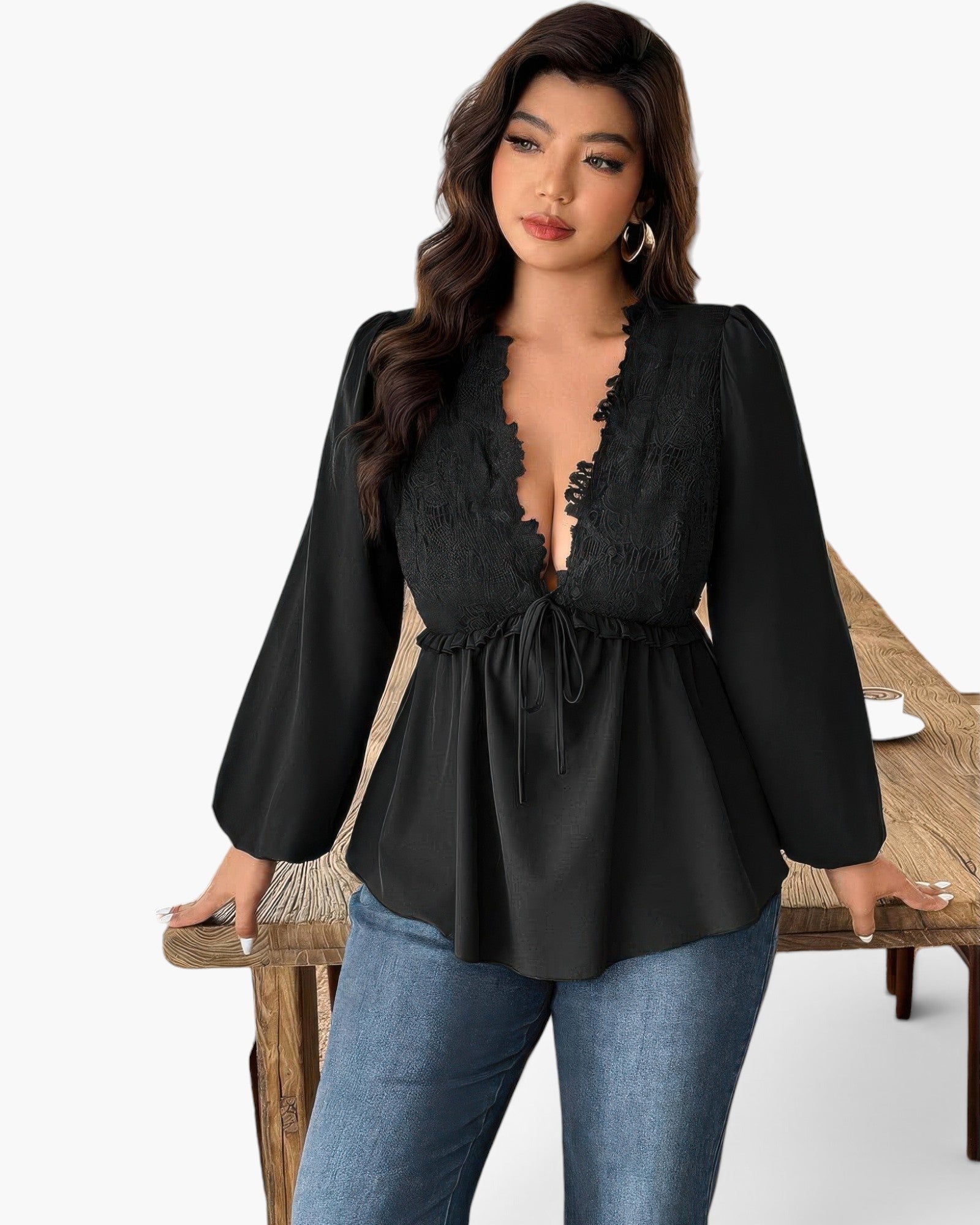 A woman modeling a black long-sleeve blouse with a lace-trimmed deep V-neckline and drawstring waist, paired with blue jeans, standing beside a rustic wooden table against a white studio background.
