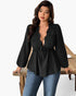 A woman modeling a black long-sleeve blouse with a lace-trimmed deep V-neckline and drawstring waist, paired with blue jeans, standing beside a rustic wooden table against a white studio background.