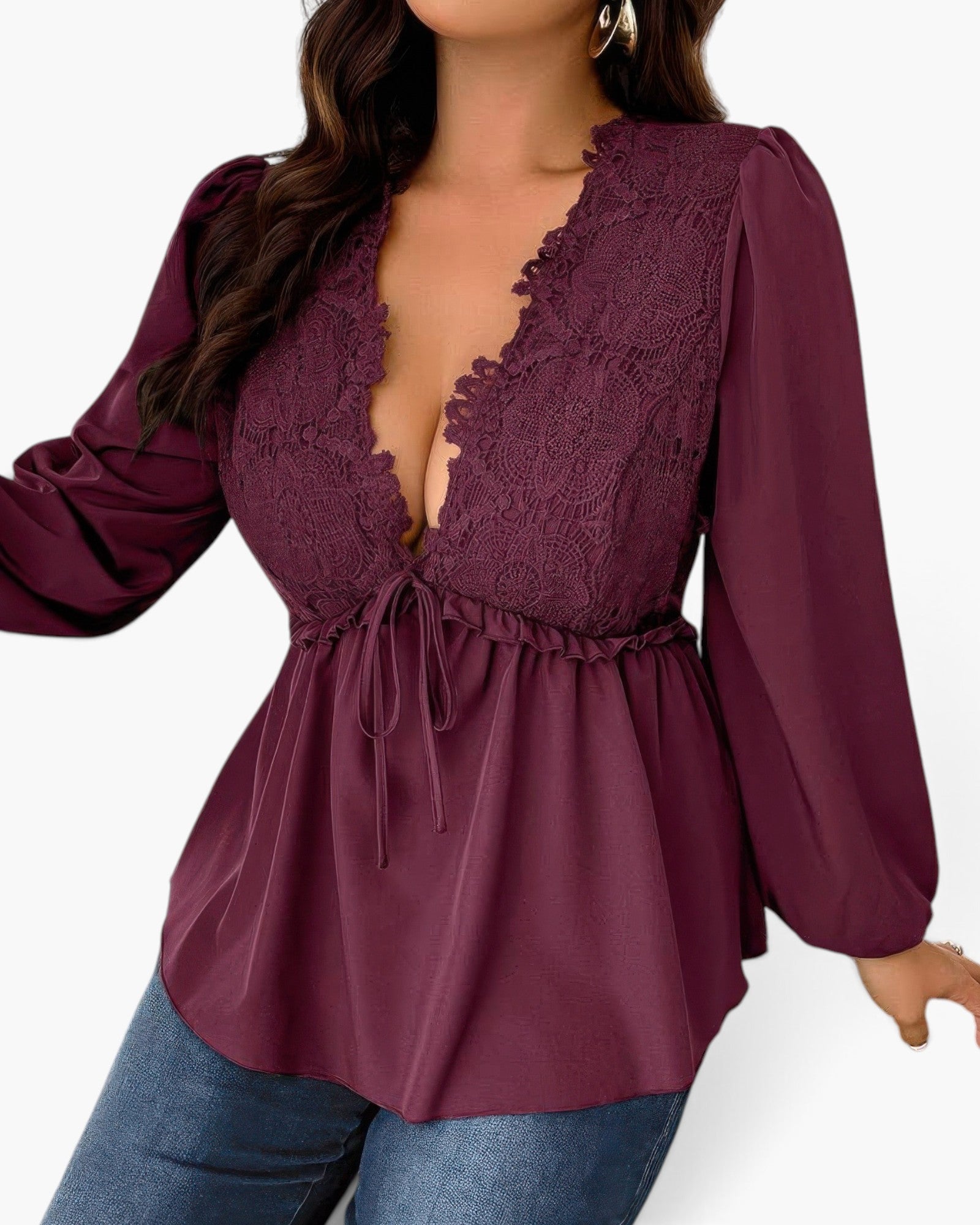 A woman wearing a long-sleeve deep burgundy blouse with a lace V-neck bodice, tie waist, and gathered peplum hem, paired with blue jeans against a light gray background.