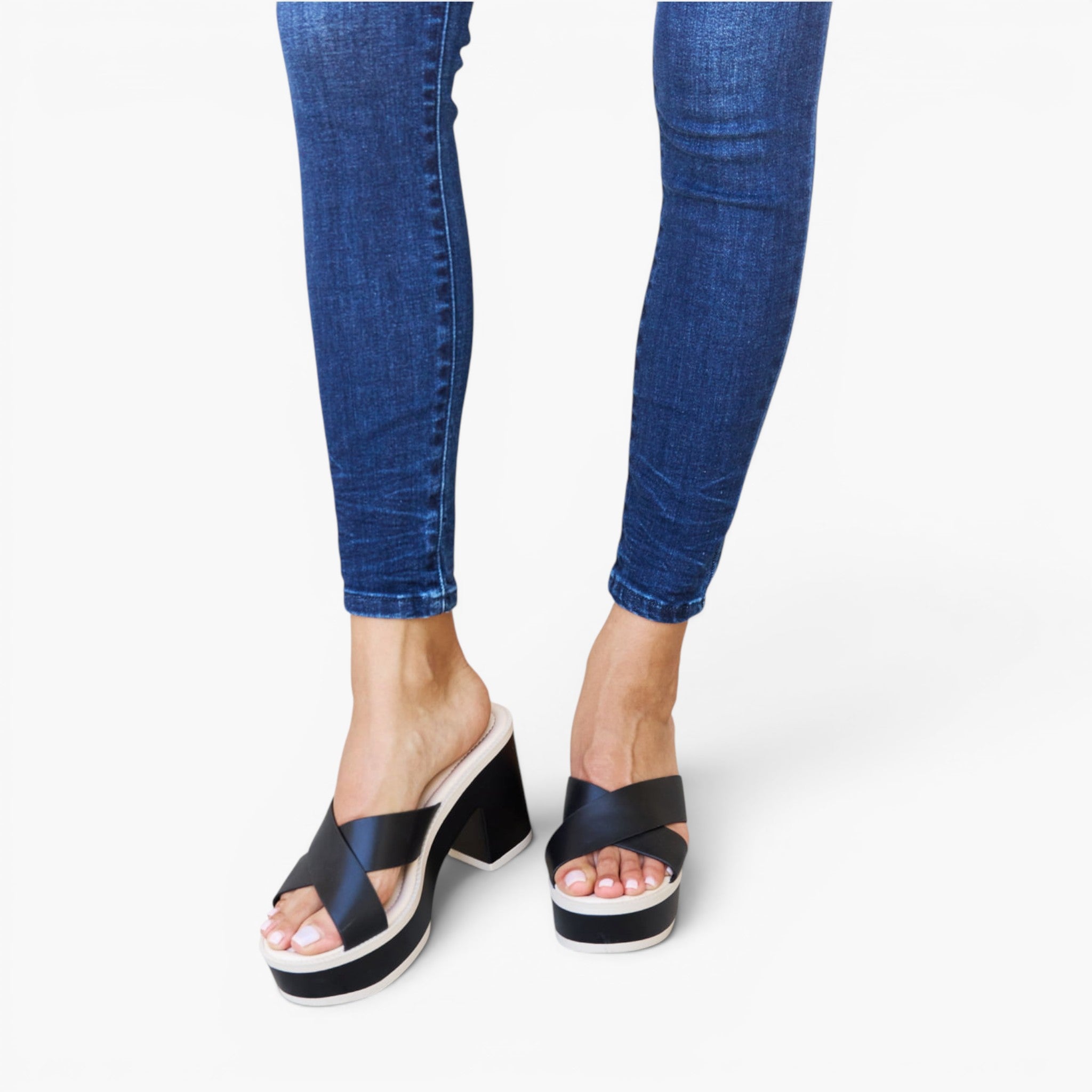 A person wearing blue skinny jeans and black platform slide sandals with crisscross straps, shown from the calves down on a clean white studio background.