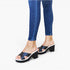 A person wearing blue skinny jeans and black platform mule sandals with white soles, shown from the calves down against a plain white studio background.