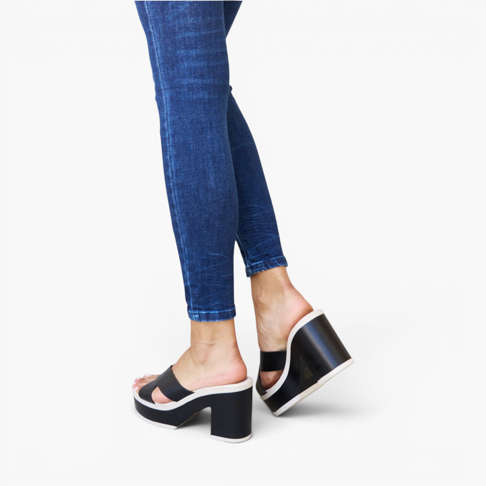 A person wearing blue skinny jeans and black platform mule sandals with white trim, shown from the calves down against a clean white studio background.