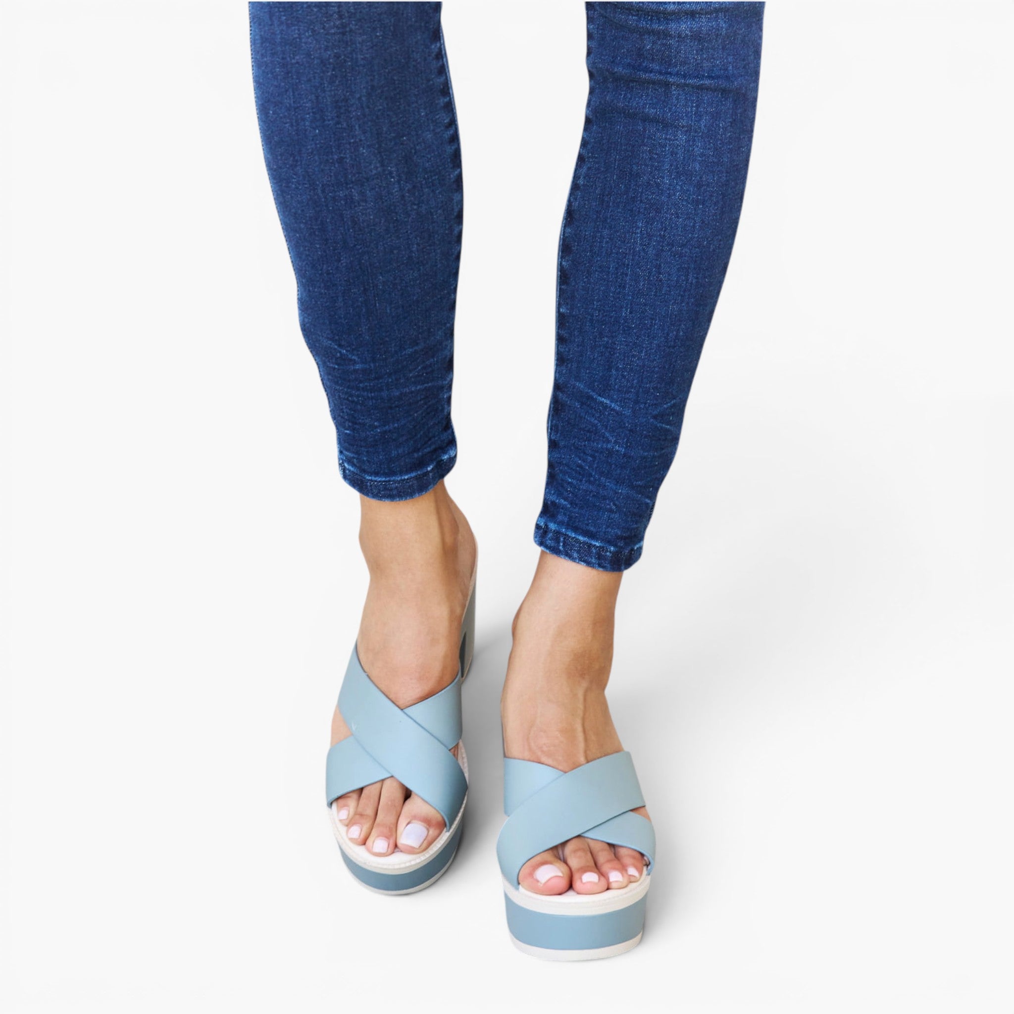 A person wearing blue skinny jeans and light blue platform slide sandals with crisscross straps, shown from the calves down on a clean white studio background.