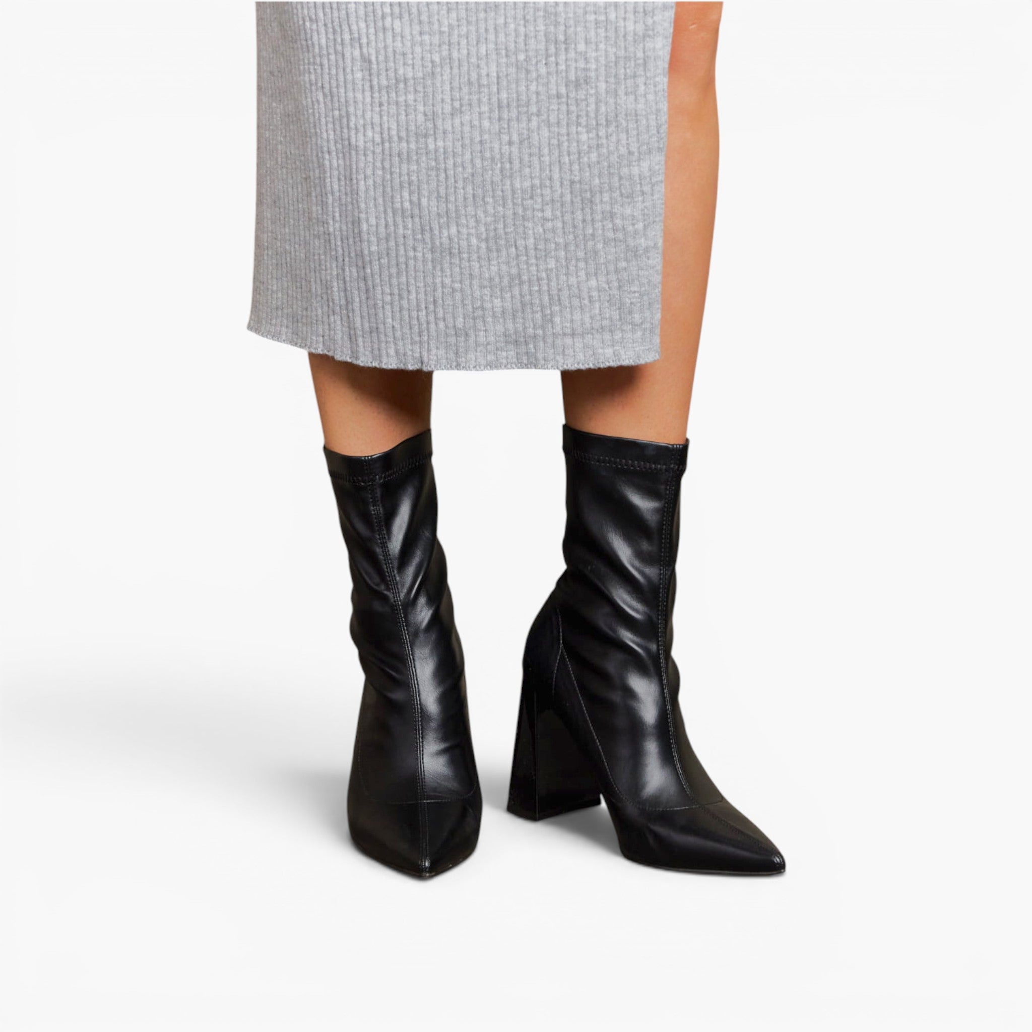 Black pointed-toe ankle boots with a high block heel, worn with a light gray ribbed skirt against a plain white background.