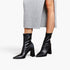 Black pointed-toe ankle boots with block heels, shown on a model wearing a light gray ribbed skirt against a plain white studio background.