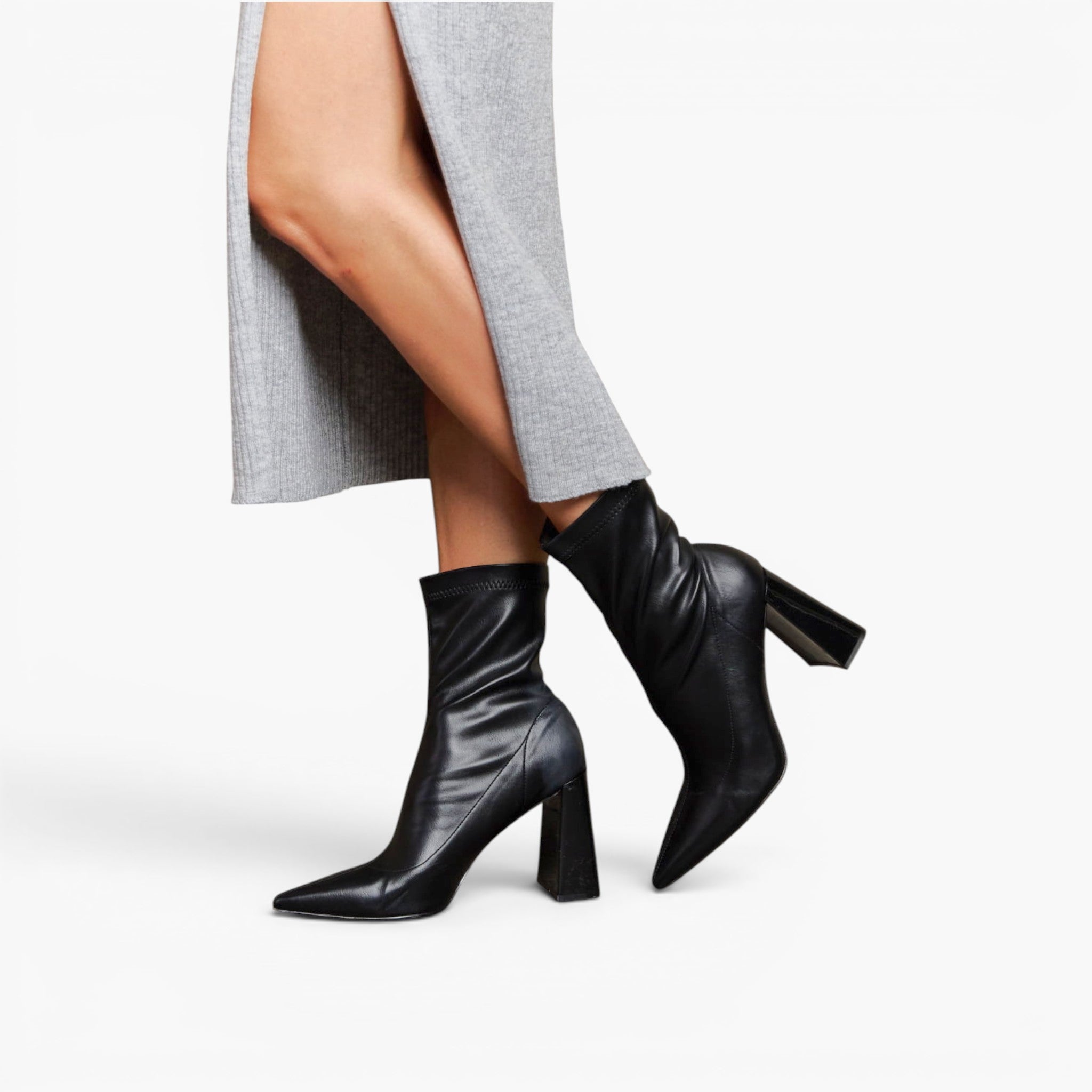 Black leather ankle boots with pointed toes and block heels, shown on a model wearing a gray draped dress against a light gray studio background.