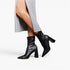 Black leather ankle boots with pointed toes and block heels, shown on a model wearing a gray draped dress against a light gray studio background.