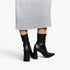 Black leather ankle boots with block heels and pointed toes, shown on a model wearing a gray ribbed midi skirt against a light gray studio background.