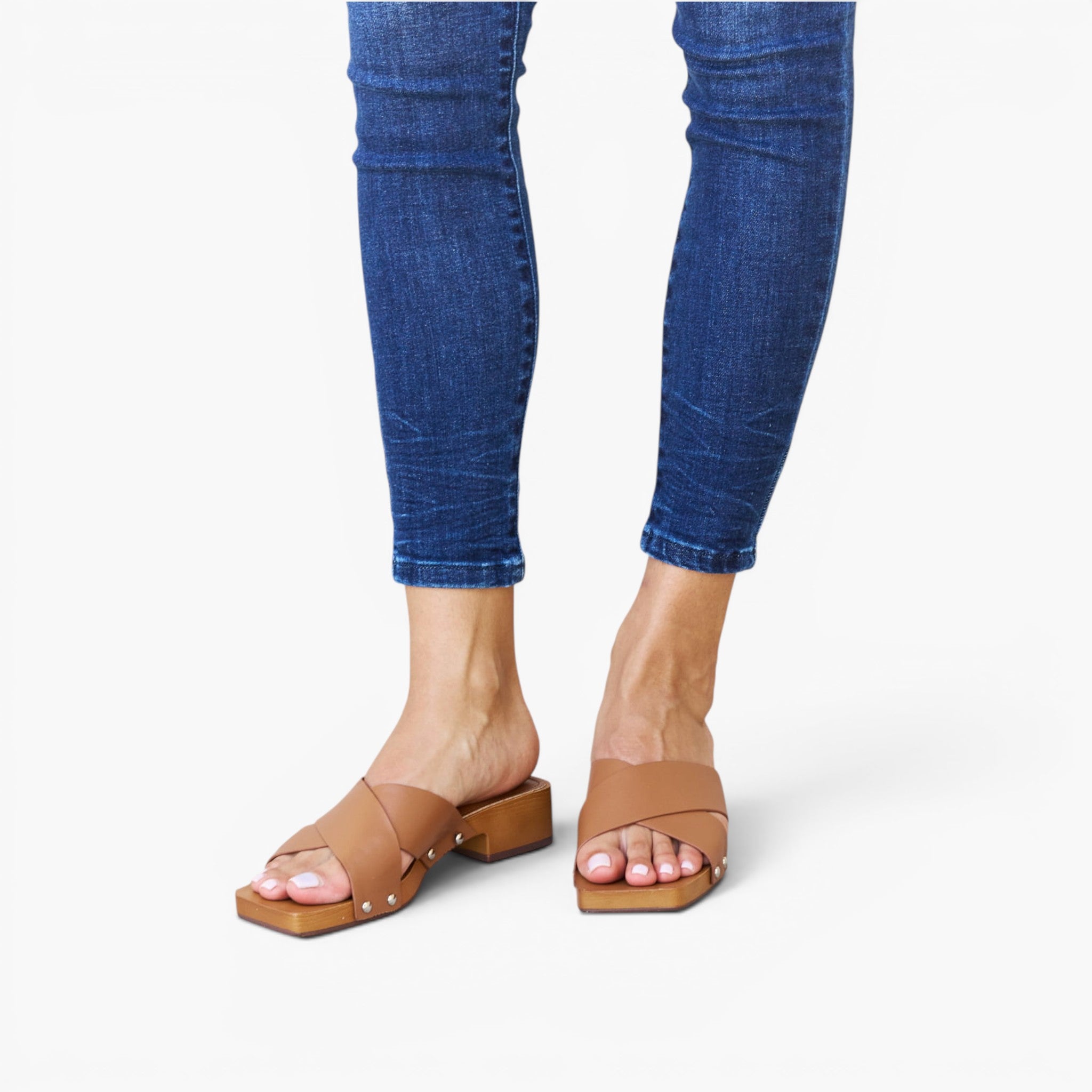 A person wearing blue skinny jeans and tan leather slide sandals with square block heels, shown from the calves down on a light gray studio background.