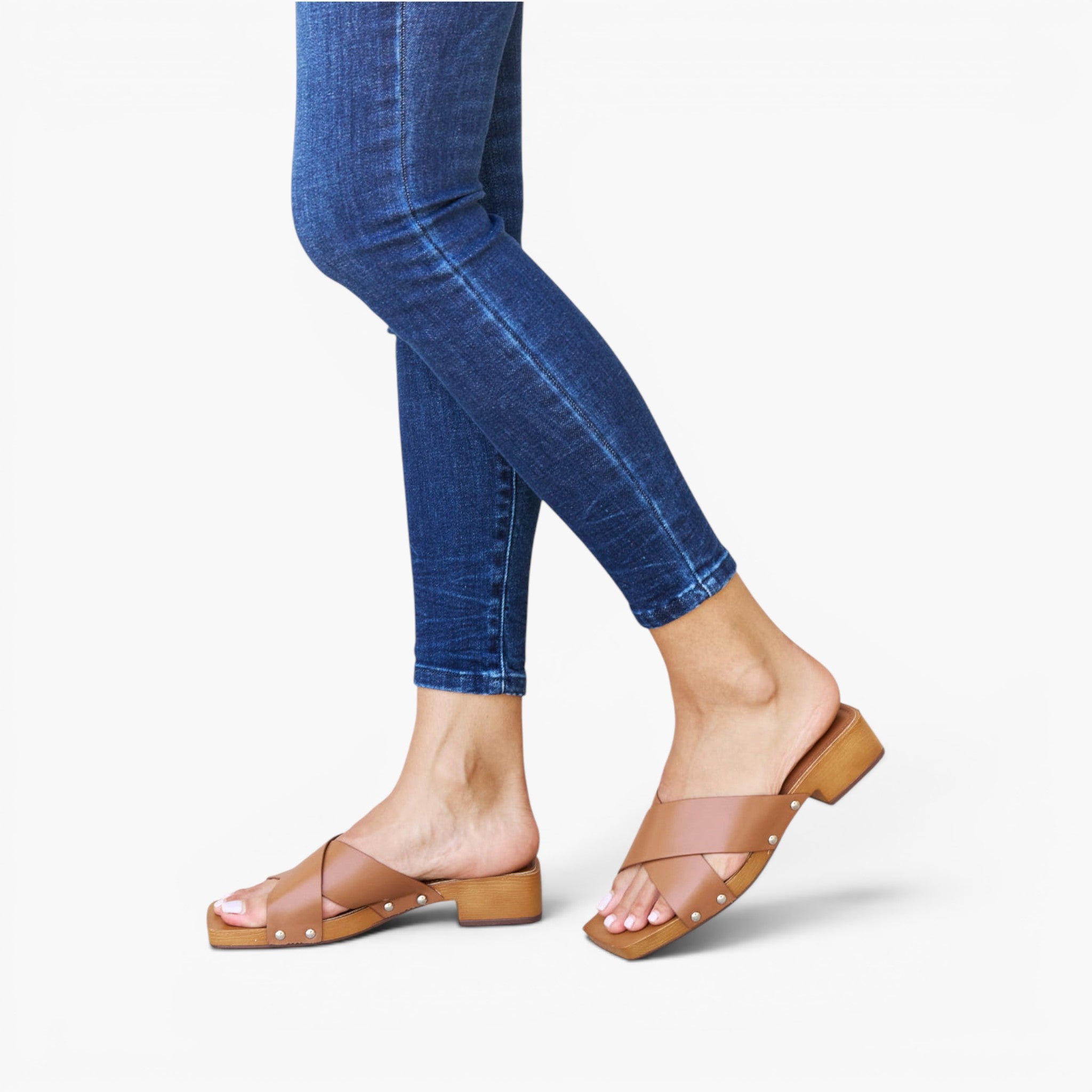 A person wearing blue skinny jeans and tan open-toe mule sandals with metallic stud detailing, shown from the calves down against a clean white studio background.