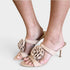 Beige strappy mule sandals with large ruffled flower embellishments on the toes, shown on a model's feet against a light gray background.
