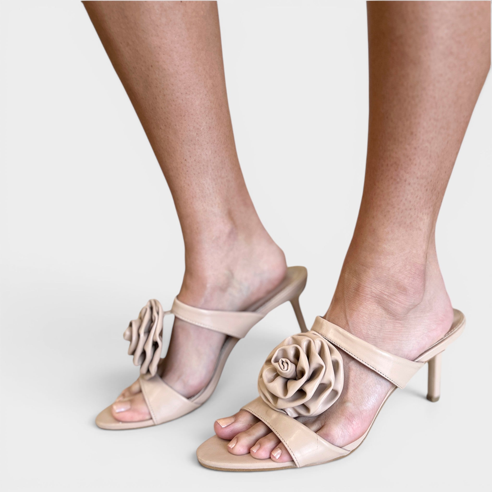 Beige satin mule sandals with a large fabric flower detail on the toe strap, shown on a model's feet against a light gray background.