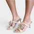 Beige satin mule sandals with a large fabric flower detail on the toe strap, shown on a model's feet against a light gray background.
