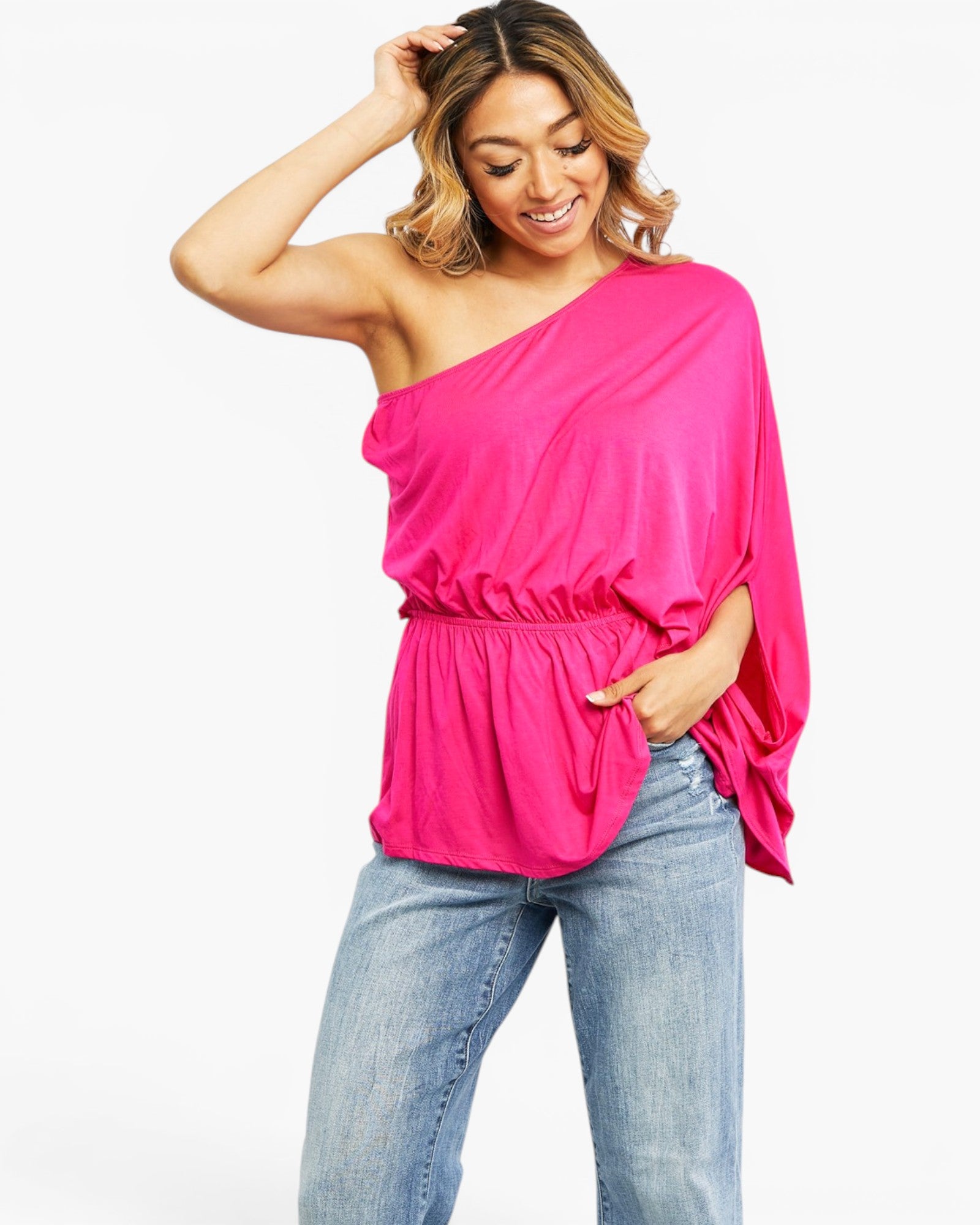 A woman modeling a bright pink one-shoulder draped top with a gathered waist, paired with light-wash blue jeans, against a plain white background.
