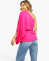A woman wearing a bright pink one-shoulder draped top and blue jeans, shown from a back-three-quarter angle against a plain white studio background.